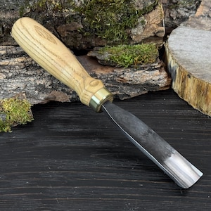 May include: A wood carving chisel with a light-colored wooden handle and a metal blade. The handle has a brass ferrule and a carved design. The blade is dark with a polished edge. The tool is designed for woodworking and carving.