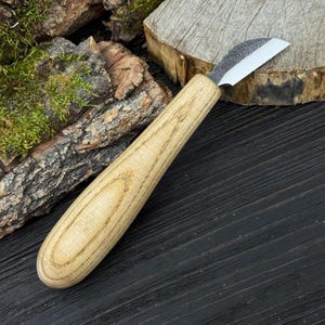 May include: A wood carving knife with a curved blade and a light-colored wooden handle. The blade is silver and the handle has a natural wood grain pattern. The knife is designed for detailed woodworking and carving projects.