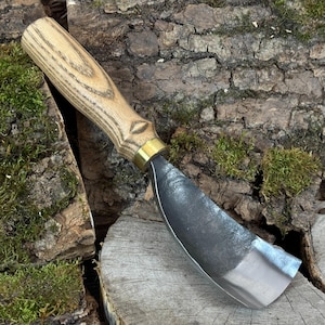 Könnte beinhalten: Ein Ziehmesser für die Holzbearbeitung mit einem hellen Holzgriff und einer gebogenen, silberfarbenen Klinge. Der Griff hat eine Messingzwinge. Das Werkzeug dient zum Formen von Holz und wird auf einer Holzoberfläche mit Moos gezeigt.