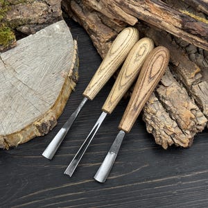 May include: Three wood carving tools with wooden handles and metal blades. The handles have a light brown wood grain pattern. The blades are silver and vary in shape, including a V-shaped gouge and a curved chisel. The tools are arranged on a dark wooden surface.