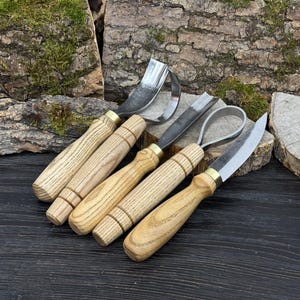 May include: A collection of wood carving tools with wooden handles and metal blades. The tools include a hook knife, a straight gouge, a curved gouge, and a detail knife. The handles are light-colored wood with brass ferrules.