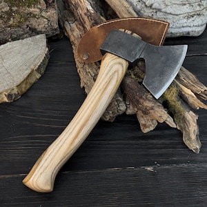 Peut inclure: Une petite hache avec un manche en bois clair et une tête en métal sombre. La lame de la hache est tranchante et a un bord incurvé. Un étui en cuir marron protège la lame. La hache est conçue pour couper du bois.