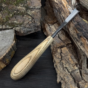 May include: A wood carving chisel with a light-colored wooden handle and a silver blade. The handle has a rounded, ergonomic shape. The chisel is designed for detailed woodworking and carving projects. The background features pieces of wood.