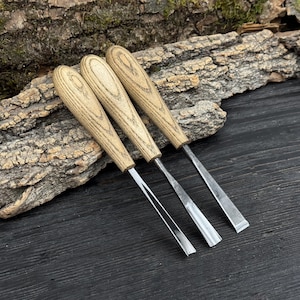 May include: Three wood carving chisels with light brown wooden handles and silver blades. The chisels are arranged diagonally on a dark wooden surface, with a piece of bark in the background. The tools are designed for woodworking and carving.