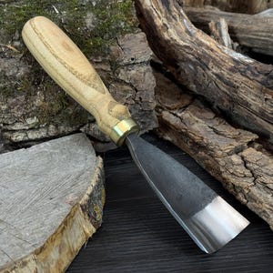 May include: A woodworking chisel with a light-colored wooden handle and a metal blade. The blade has a sharp, angled edge. The handle is attached to the blade with a brass ferrule. The chisel is designed for shaping and carving wood.