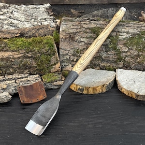 Op de afbeelding: Een houtbeitel met een lichtgekleurde houten handgreep en een donker metalen blad. Het blad heeft een zilveren rand. De beitel staat tegen een achtergrond van hout en een kleine leren schede.