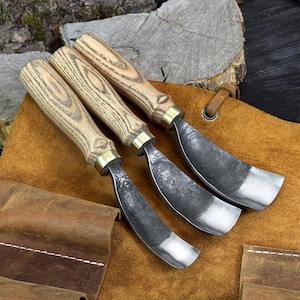 May include: Three wood carving gouges with wooden handles and metal blades. The handles are light-colored with a wood grain pattern, and the blades have a curved shape with a silver edge. The tools are arranged on a brown leather surface.