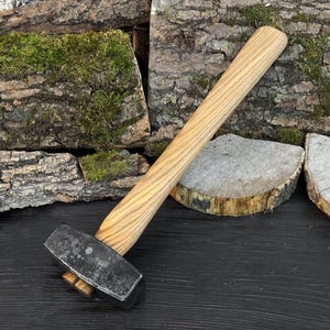 May include: A heavy-duty hammer with a light-colored wooden handle and a dark, rectangular head. The hammer is angled across a dark surface, with a backdrop of wood and moss.