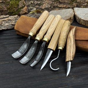 May include: A set of six wood carving tools with wooden handles and metal blades. The blades have various shapes, including curved and pointed designs. A brown leather case is in the background.