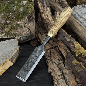 May include: A woodworking chisel with a light wood handle, brass ferrule, and a dark, textured blade. The chisel is set against a backdrop of wood and bark.