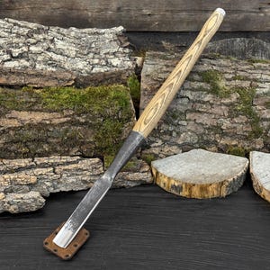 Puede incluir: Un formón para carpintería con un mango de madera clara y una hoja de metal. La hoja está fijada en una férula de metal oscuro. El formón descansa sobre un pequeño soporte cuadrado de cuero y metal marrón. El fondo presenta madera y musgo.