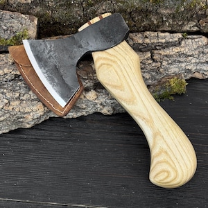 Peut inclure: Une hache artisanale avec une lame en acier sombre et une poignée en bois clair. La hache est livrée avec un étui en cuir. La poignée mesure environ 30 cm de long. Parfait pour les activités de plein air.