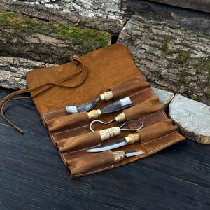 May include: A set of wood carving tools with wooden handles and metal blades, nestled in a brown leather roll. The roll is secured with a leather cord. The tools include various blade shapes for detailed carving.