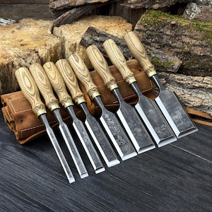 May include: A collection of seven wood chisels, each with a light wood handle and a metal blade. The chisels are displayed in a row, with a leather tool roll on the left. The blades have a dark, textured finish.