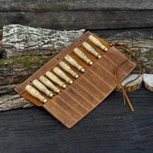 May include: A collection of wood carving chisels with light-coloured wooden handles and metal ferrules, presented in a brown leather tool roll. The roll is secured with a leather strap and has white stitching.