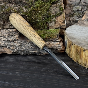 Könnte beinhalten: Ein Holzbeitel mit einem hellen Holzgriff und einer Stahlklinge. Der Griff hat eine abgerundete Form und eine strukturierte Oberfläche. Die Klinge ist rechteckig und verjüngt sich zu einer scharfen Kante. Der Stecheisen ist zum Schnitzen und Formen von Holz bestimmt.