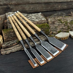 May include: Five woodworking chisels with wooden handles and metal blades are displayed. The chisels are arranged diagonally, resting on small, square leather pads. The handles have a light wood grain pattern, and the blades are silver.