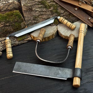 May include: A collection of handcrafted metal blades with wooden handles. The set includes a large, straight blade, two curved blades, and a cleaver-style blade. The handles are made of light-colored wood with dark bands. The blades are displayed on a dark wooden surface.