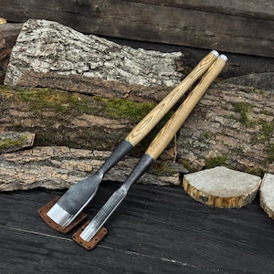 Puede incluir: Dos formones de madera con mangos de madera marrón claro y hojas de metal oscuro. Los formones descansan sobre pequeñas almohadillas cuadradas de cuero. El fondo presenta una superficie de madera y un tronco.