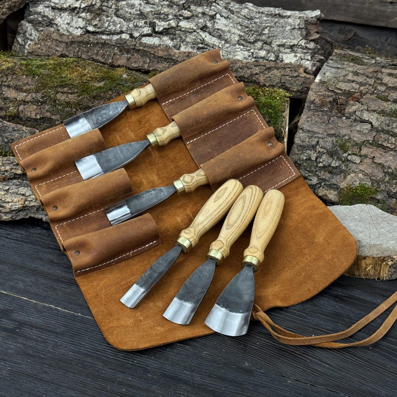 May include: A set of wood carving chisels with wooden handles and metal blades, displayed in a brown leather tool roll. The chisels are of varying sizes and shapes, designed for detailed woodworking. The leather roll has individual pockets for each chisel.