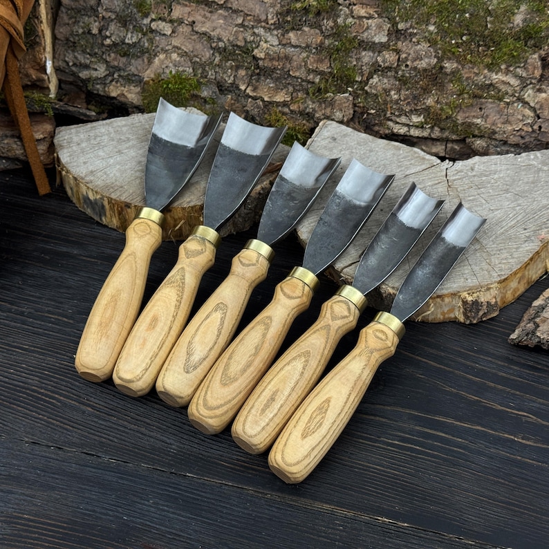 May include: Set of six wood carving gouges with light wood handles and metal blades. The blades are curved and the handles are attached with brass ferrules. The tools are arranged on a dark wooden surface.