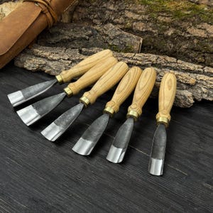 May include: Set of six wood carving chisels with wooden handles and metal blades. The chisels are arranged in a row, with the blades facing the same direction. The handles are light-colored wood, and the blades are silver.