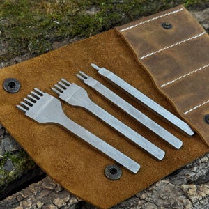 Peut inclure: Un ensemble de quatre outils de travail du cuir argentés présentés sur une pochette en cuir marron. Les outils comprennent un poinçon à plusieurs dents, un poinçon à deux dents et deux autres outils. La pochette en cuir est ouverte, révélant les outils.