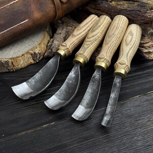 May include: Four wood carving gouges with curved blades. The gouges have light-coloured wooden handles with brass ferrules. The blades are made of dark metal. The tools are arranged on a dark wooden surface, with a leather sheath in the background.