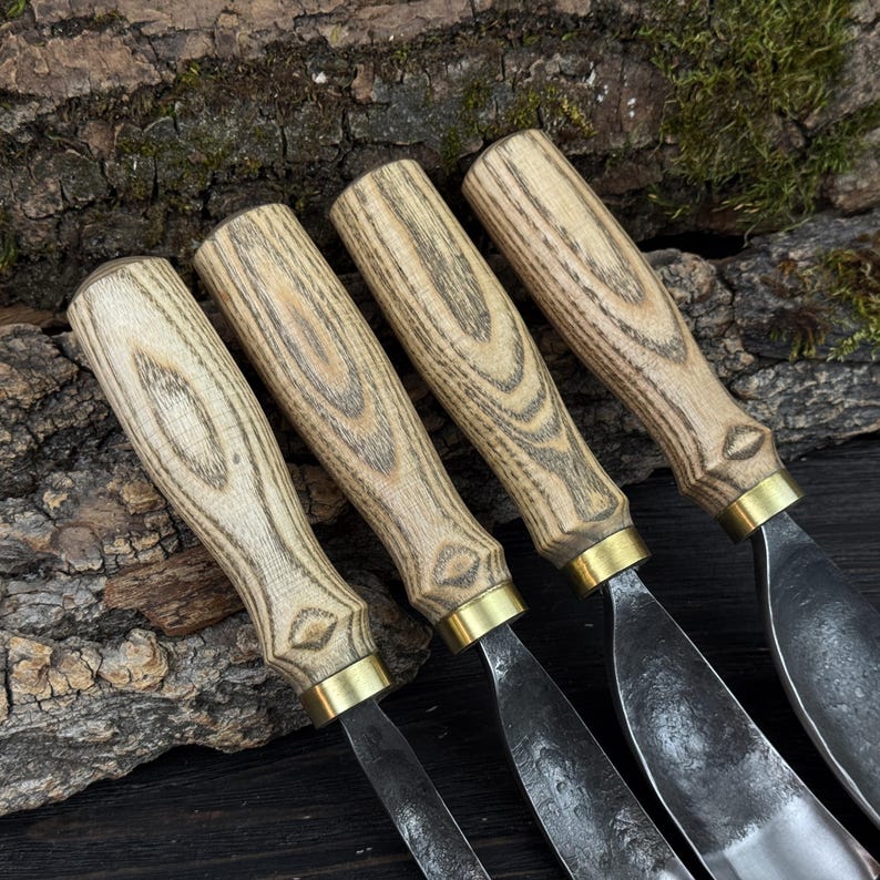 May include: Four wood carving tools with light-coloured wooden handles and metal blades. The handles have a natural wood grain pattern and are accented with brass collars. The blades are a dark, matte metal. The tools are arranged on a textured, dark wood surface.
