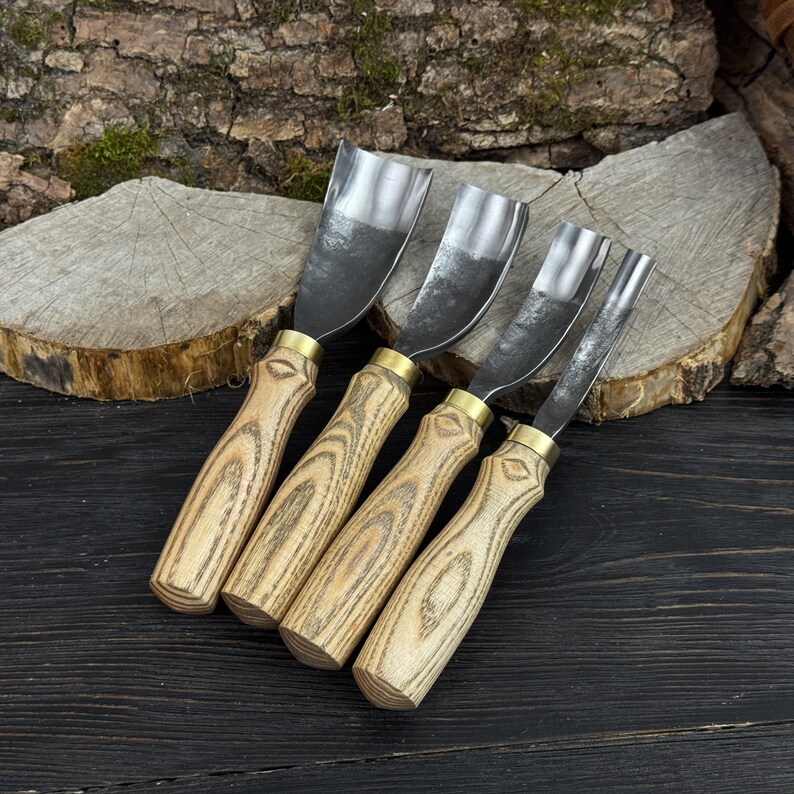 May include: Four wood carving gouges with curved steel blades. The handles are made of light-coloured wood with a natural grain pattern, and are connected to the blades with brass ferrules. The tools are arranged on a dark wooden surface.