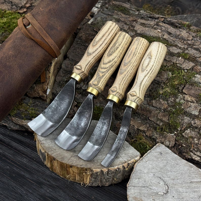 May include: Four wood carving gouges with wooden handles and metal blades. The gouges are displayed on a wooden surface, with a leather tool roll in the background. The blades have a curved shape.