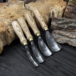 May include: Four wood carving gouges with curved blades. The handles are light-coloured wood with a brass ferrule. The blades are made of dark metal. The tools are arranged on a dark wooden surface with pieces of bark in the background.