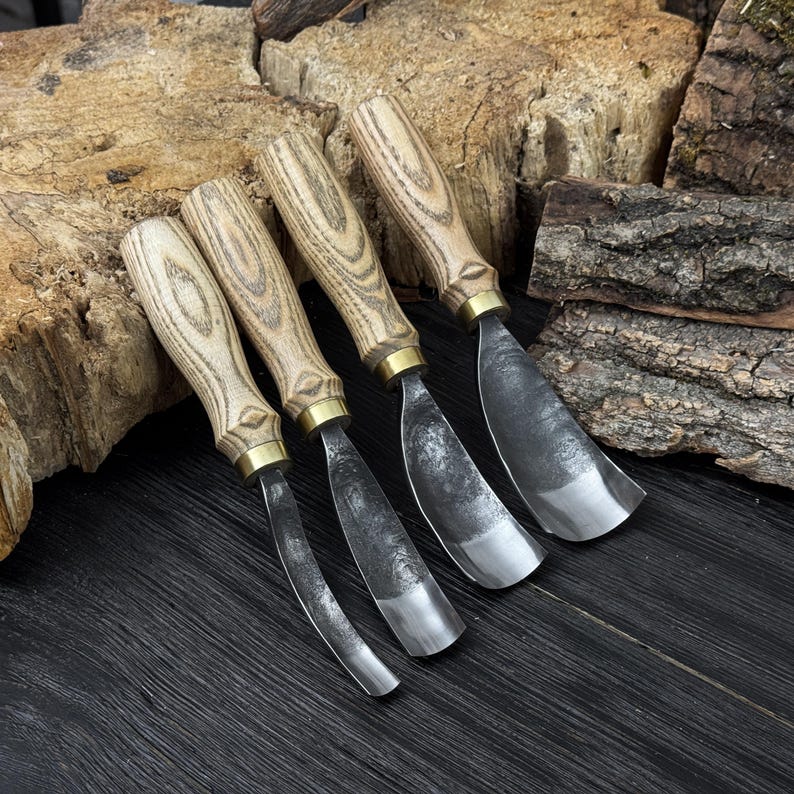 May include: Four wood carving gouges with curved blades. The handles are light-coloured wood with a brass ferrule. The blades are made of dark metal. The tools are arranged on a dark wooden surface with pieces of bark in the background.