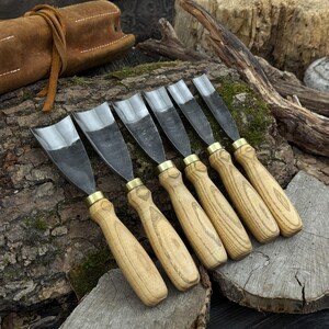 May include: A set of six wood carving gouges with polished steel blades and light wood handles. The gouges are arranged in a row on a wooden surface, with a leather tool roll in the background. The blades have a curved shape.