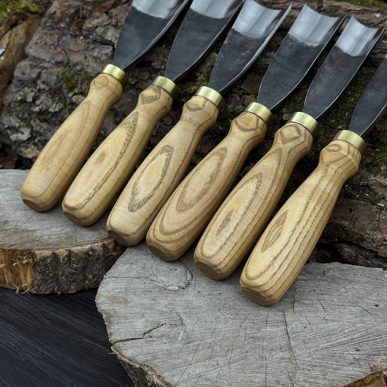 May include: Six wood carving gouges with light brown wooden handles and curved steel blades. The handles have a natural wood grain pattern and are connected to the blades with brass ferrules. The tools are arranged on a rustic wooden surface.