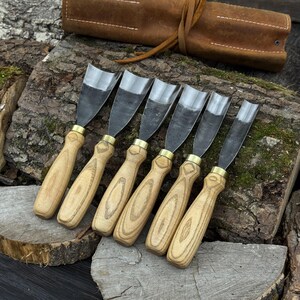 May include: A set of six wood carving chisels with polished steel blades and light-colored wooden handles. The chisels are arranged in a row, with a brown leather tool roll in the background.