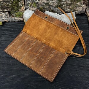 Può includere: Astuccio arrotolabile in pelle marrone aperto, con più scomparti, assicurato da un laccio in pelle. La custodia presenta cuciture bianche e occhielli metallici. L'interno è di una tonalità marrone più chiara.
