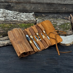 May include: A tan leather tool roll containing five wood carving tools with light-colored wooden handles and metal blades. The roll features individual slots for each tool and a long leather tie for securing the wrap when it is rolled closed.