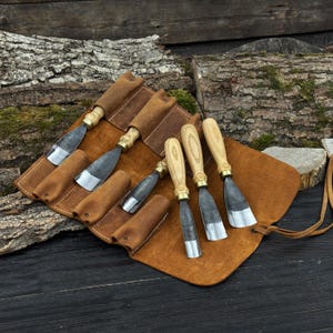 May include: A set of wood carving chisels with wooden handles and metal blades, displayed in a brown leather tool roll. The chisels have brass ferrules and are arranged on a rustic wooden surface.