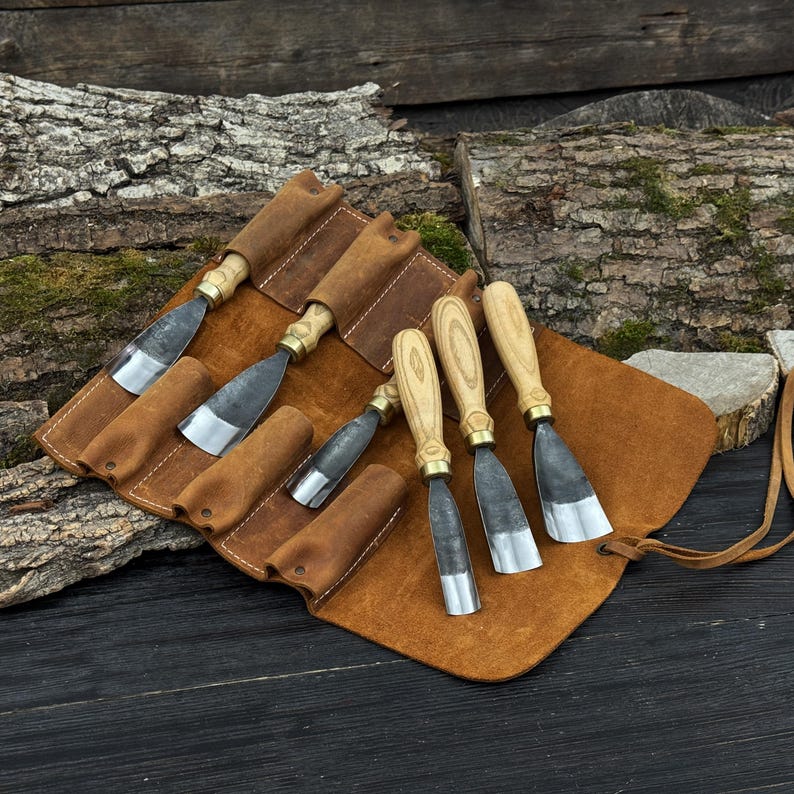 May include: A set of wood carving chisels with wooden handles and metal blades, displayed in a brown leather tool roll. The chisels have brass ferrules and are arranged on a rustic wooden surface.