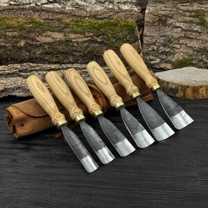 May include: A set of seven wood carving chisels with light-colored wooden handles and metal blades. The chisels are arranged in a row, with a brown leather tool roll on the left. The blades have a silver finish.