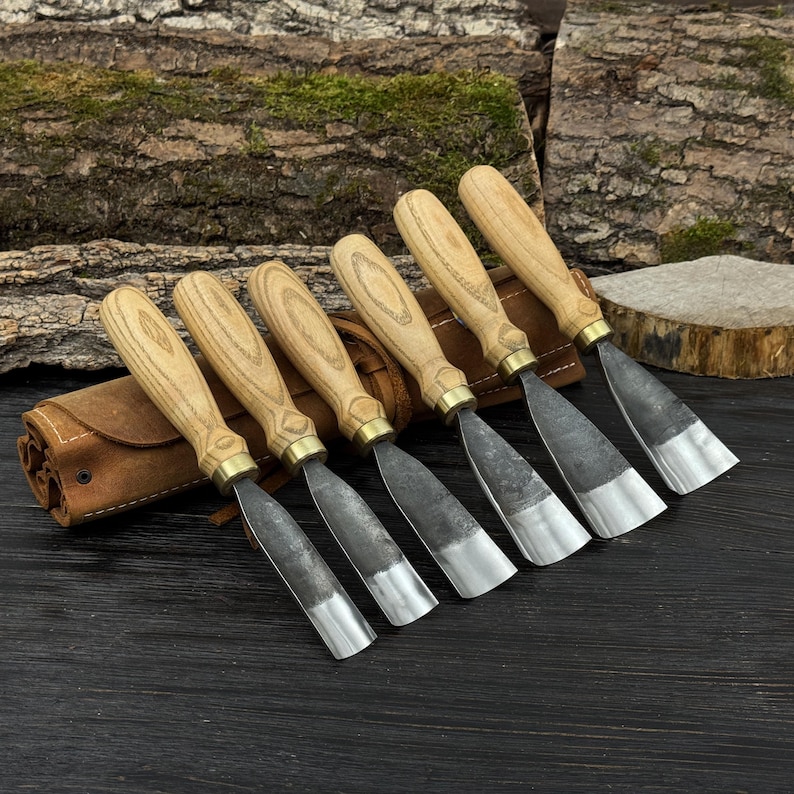 May include: A set of seven wood carving chisels with light-colored wooden handles and metal blades. The chisels are arranged in a row, with a brown leather tool roll on the left. The blades have a silver finish.