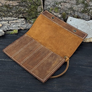 May include: Open brown leather roll-up case. The case has three interior pockets with white stitching and a tan interior. A leather strap with a knot closure is visible.