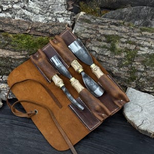 May include: Four wood carving tools with metal blades and wooden handles, presented in a brown leather tool roll. The handles feature brass detailing. The tool roll is open, displaying the tools. The background is a wooden surface.