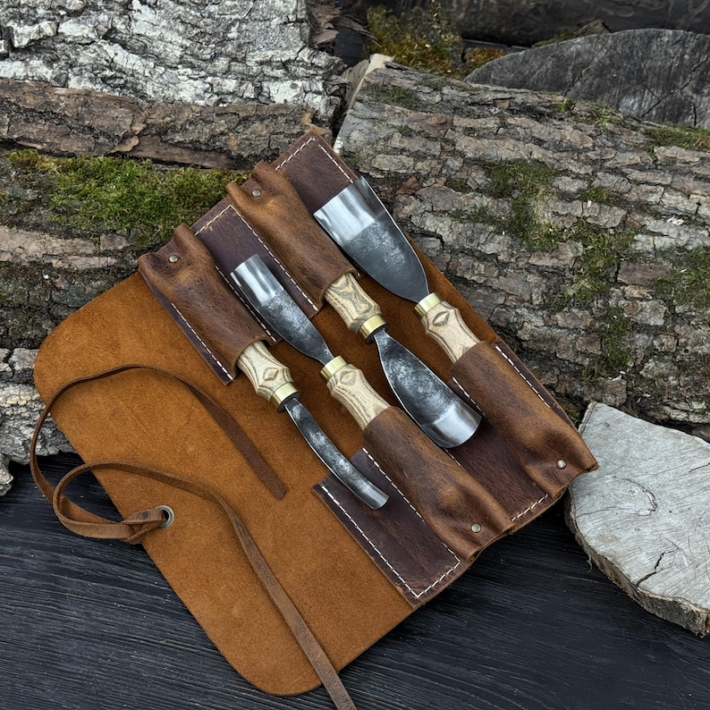 May include: Four wood carving tools with metal blades and wooden handles, presented in a brown leather tool roll. The handles feature brass detailing. The tool roll is open, displaying the tools. The background is a wooden surface.