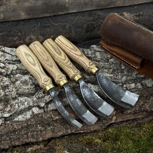 May include: Four wood carving gouges with wooden handles and metal blades. The handles are light-coloured wood with a natural grain pattern, and the blades are curved. A brown leather sheath is visible in the background.