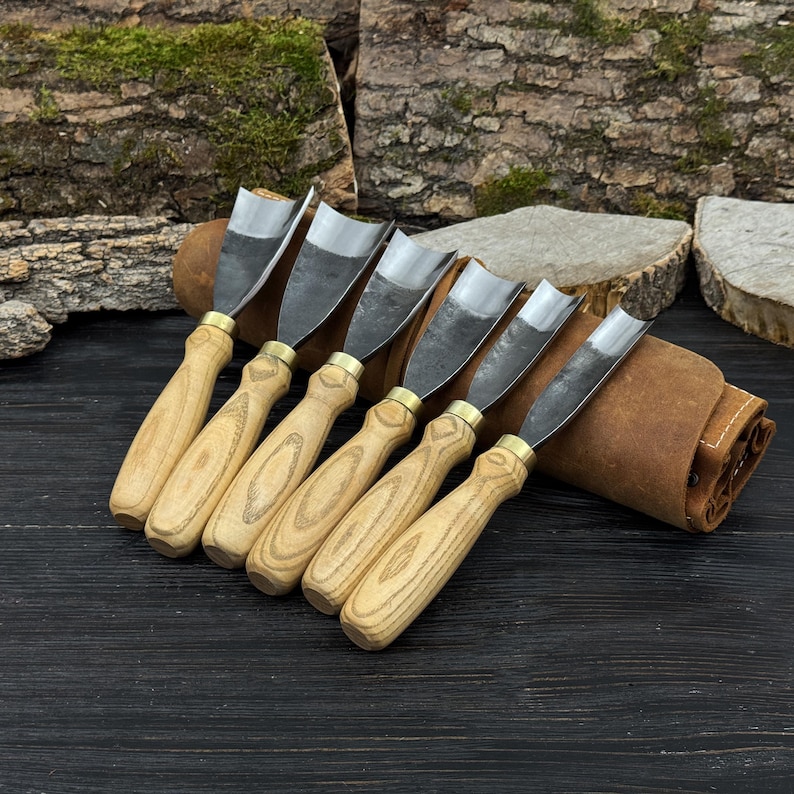 May include: A set of six wood carving gouges with wooden handles and metal blades. The tools are arranged in a row, with a leather tool roll in the background. The handles are light-colored wood, and the blades are silver.
