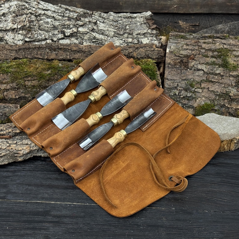 May include: A set of six wood chisels with wooden handles and metal blades, displayed in a brown leather tool roll. The chisels are arranged in a row, with the blades visible. The tool roll is open, revealing the tools. The background features wood.