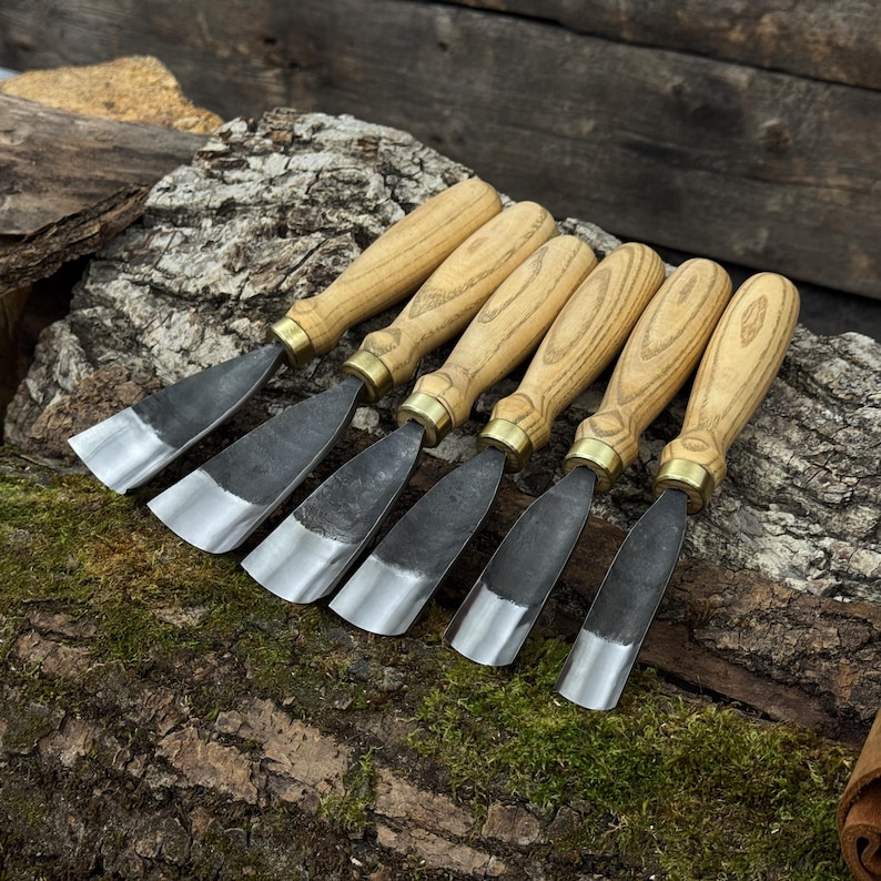 May include: Six wood carving chisels with light-colored wooden handles and metal blades. The blades have a dark, textured finish with a polished, silver edge. The chisels are arranged on a mossy, textured surface.