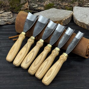 May include: A set of six wood carving gouges with polished steel blades and light-colored wooden handles. The handles have a natural wood grain pattern and are connected to the blades with brass ferrules. The tools are arranged in a row, suggesting a woodworking or carving set.